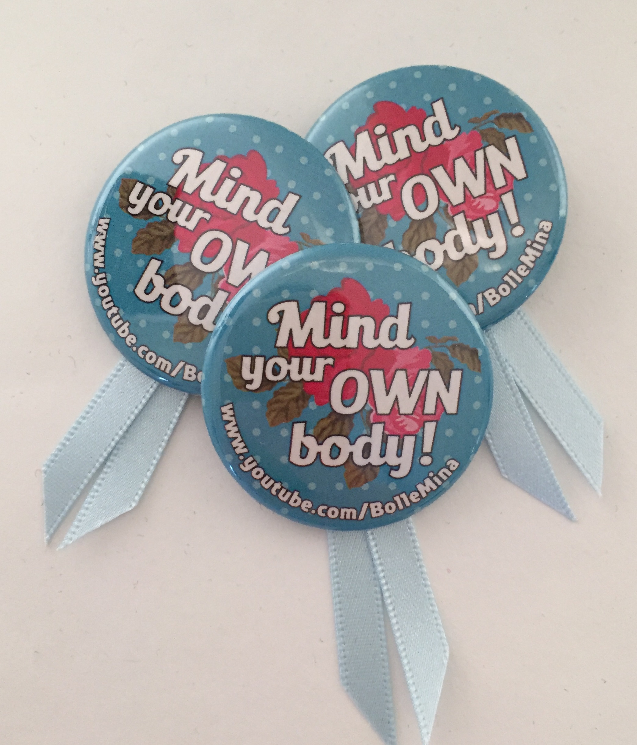 Foto van drie buttons met de slogan Mind your own body erop. Met lichtblauwe satijnen lintjes eronder geplakt. Dit is het symbool van International No Diet Day